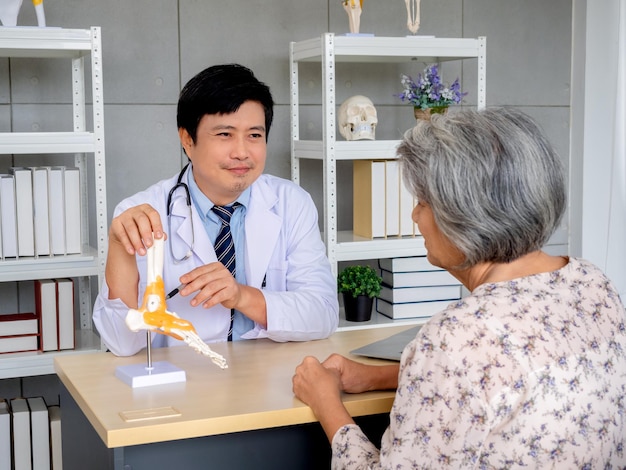 Médico ortopédico profesional asiático sonriente que señala el modelo de anatomía de la articulación del tobillo del esqueleto del pie para explicar a una paciente mayor en el consultorio médico Concepto de atención médica de tratamiento fisiológico