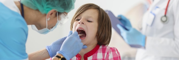 Médico otorrinolaringologista em máscara médica protetora, examinando a garganta de uma menina usando uma espátula.