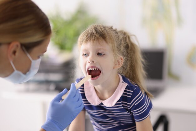 Médico otorrinolaringólogo en máscara médica protectora examinando la garganta de la niña con una espátula de madera