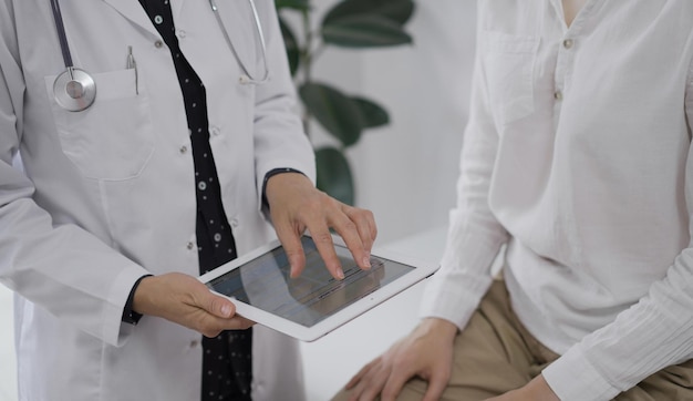 El médico y un paciente discutiendo algo La médica está señalando a la pantalla de la tableta con notas médicas además de una mujer joven durante una consulta en la clínica