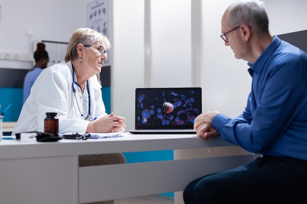 Médico y paciente discutiendo sobre la ilustración del coronavirus en la computadora portátil. Médico general que usa computadora para explicar la representación visual de la bacteria covid 19 al hombre mayor.