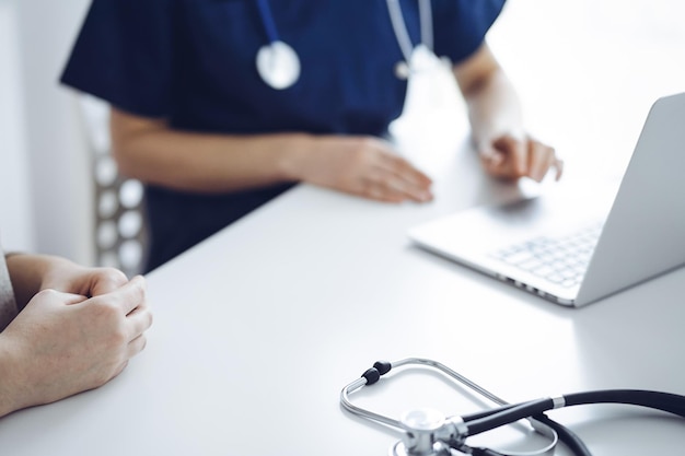 Médico y paciente hablando entre sí durante el examen de salud actual mientras están sentados en el escritorio en la clínica, de cerca de la mano.