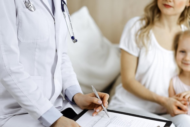 Médico y paciente. Pediatra usando portapapeles mientras examina a una niña con su madre en casa. Niño caucásico lindo feliz en el examen médico. Concepto de medicina.