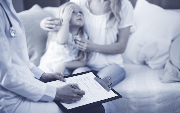 Médico y paciente. Pediatra usando portapapeles mientras examina a una niña con su madre en casa. Niño enfermo e infeliz en el examen médico. Concepto de medicina.