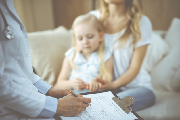 Médico y paciente. Pediatra usando portapapeles mientras examina a una niña con su madre en casa. Niño enfermo e infeliz en el examen médico. Concepto de medicina.