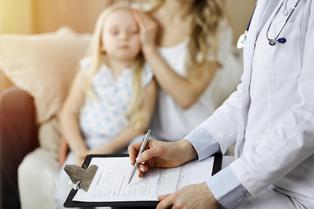 Médico y paciente. Pediatra usando portapapeles mientras examina a una niña con su madre en casa. Niño enfermo e infeliz en el examen médico.