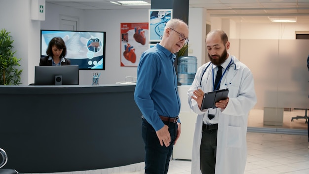 Médico y paciente senior mirando tableta digital en el vestíbulo de recepción del hospital, hablando de diagnóstico médico en la sala de espera. Cita de visita de control con especialista para dar consejos.