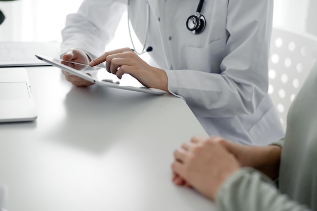 Foto el médico y el paciente sentados en el escritorio de la oficina de la clínica el enfoque está en las manos de la médica usando la tableta de computadora de cerca concepto de medicina