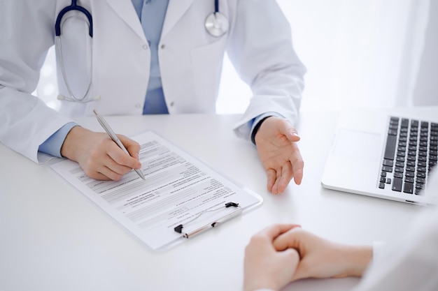 Médico y paciente sentados uno frente al otro en el escritorio de la clínica. La atención se centra en las manos de la médica que llenan el formulario de registro del historial de medicamentos o la lista de verificación, de cerca. Concepto de medicina.