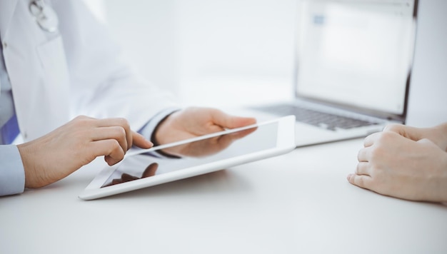 El médico y el paciente sentados uno frente al otro en el escritorio de la clínica. El enfoque está en las manos de la médica usando la tableta, de cerca. Concepto de medicina.