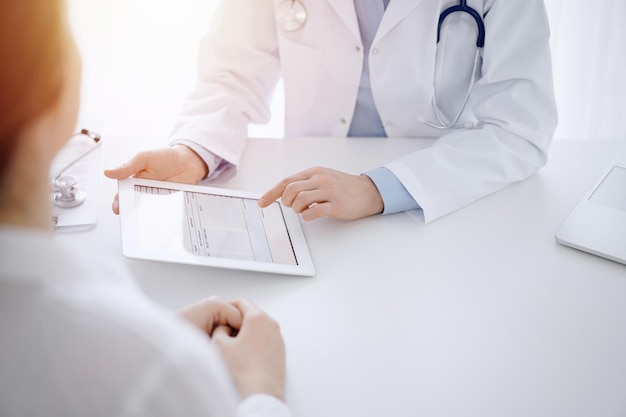 Médico y paciente sentados uno frente al otro en el escritorio de la clínica. El foco está en las manos de la doctora apuntando al panel táctil de la tableta, de cerca. concepto de medicina.