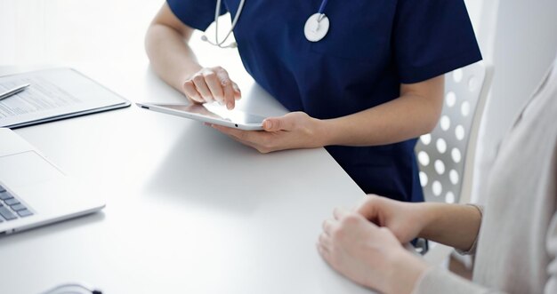 Médico y paciente sentados en la mesa de la clínica mientras usan una tableta. La atención se centra en las manos de la médica, de cerca. Concepto de medicina.