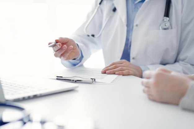 Médico y paciente sentados en la mesa en la oficina de la clínica. El foco está en la mano de la médica apuntando al monitor de la computadora portátil, de cerca. Concepto de medicina.