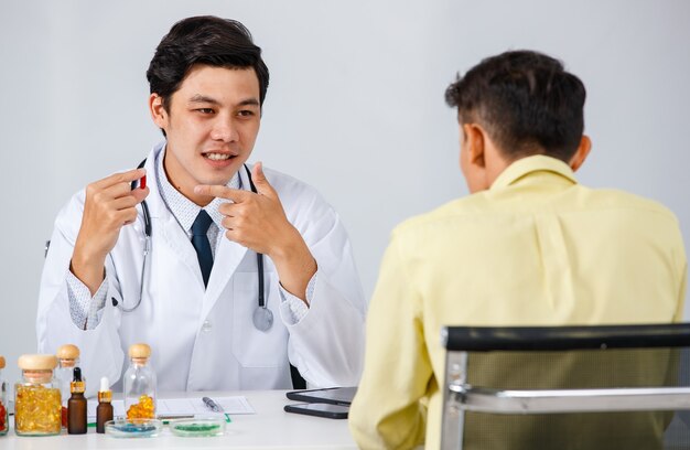 Médico y paciente de sexo masculino asiático alegre sentado en la mesa y hablando de píldoras mientras se miran durante la cita sobre fondo blanco