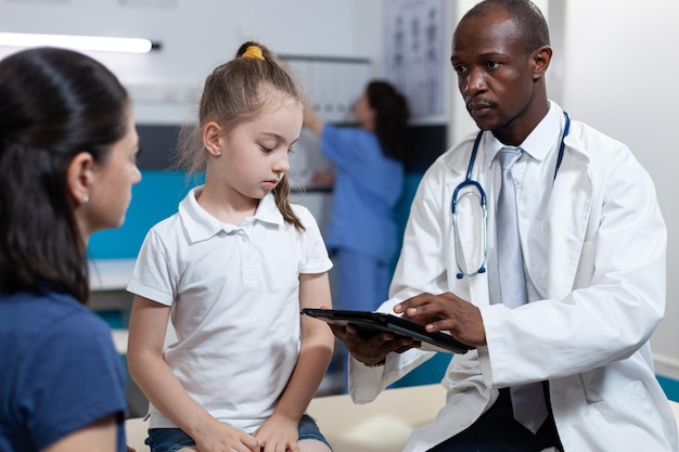 Médico pediatra afro-americano que analisa os sintomas da doença, digitando o tratamento de saúde durante a consulta médica no consultório do hospital. homem terapeuta discutindo conhecimento sobre doença com a família