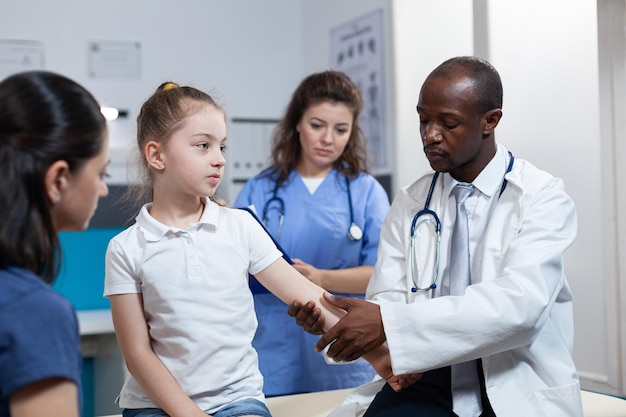 Médico pediatra afroamericano vendaje hueso fracturado de paciente niña durante fisioterapia clínica en la oficina del hospital. Niño pequeño con un brazo roto después de un accidente. Servicios de salud