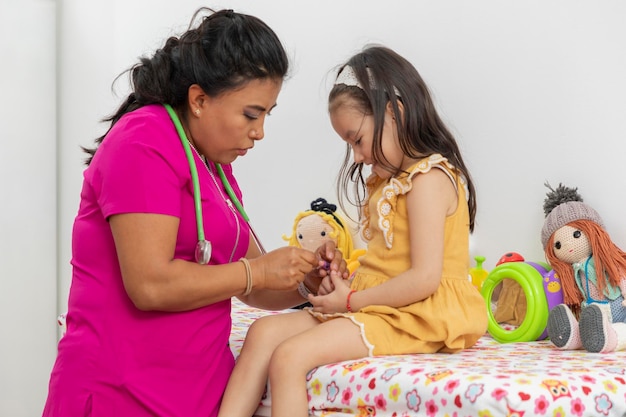 Médico pediatra colocando una tirita en el dedo de una niña con un vestido amarillo sentada en el sofá de la oficina