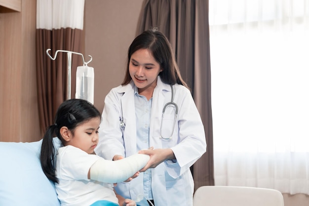 Foto médico pediatra examina a la niña asiática con un brazo roto con un yeso en el hospital