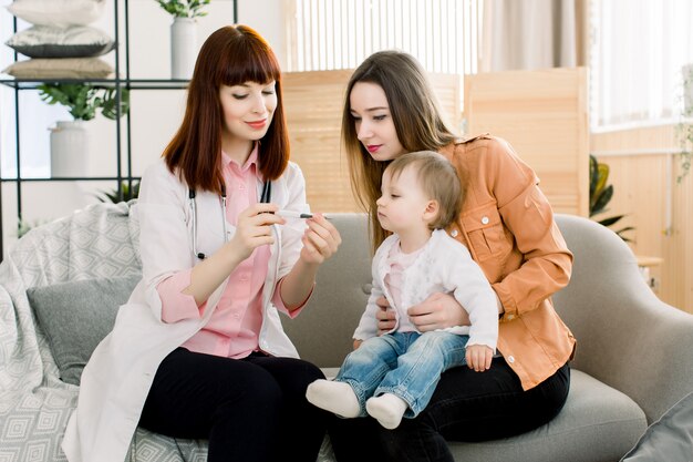 Médico pediatra, examinando a temperatura da menina nos braços da mãe. Médico infantil, verificando a temperatura do bebê no hospital