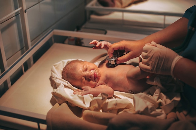 Médico pediatra examinando al bebé en la clínica, bebé recién nacido llorando en la cama