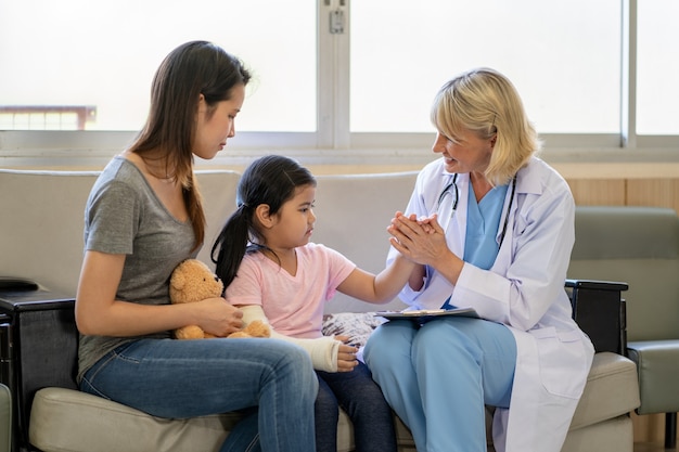 Médico pediatra examinando menina asiática com braço quebrado usando gesso no hospital