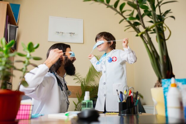 Médico pediatra examinando a un niño en el consultorio médico comfortabe. Concepto de salud, infancia, medicina, protección y prevención. El niño confía en el médico y se siente tranquilo, con máscara.