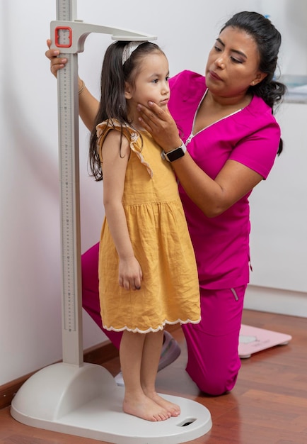 Médico pediatra latino mide a una niña en su oficina con una regla de pedestal