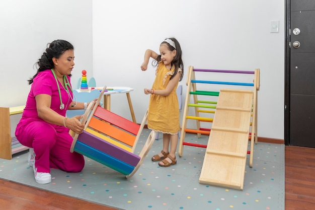 Médico pediatra latino verificando o desenvolvimento motor de uma garota na área de recreação de seu escritório