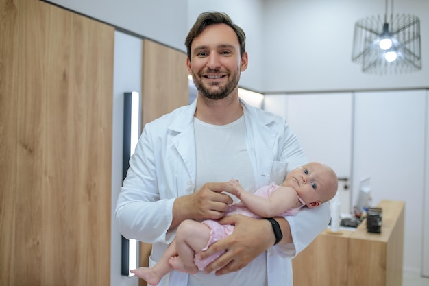 Médico pediátrico sonriente y amigable parado en el área de recepción y sosteniendo al recién nacido en sus brazos