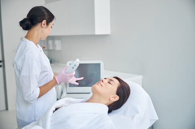 Médico de pelo oscuro con una bata de laboratorio y guantes de látex tocando la pantalla del monitor de la máquina de belleza