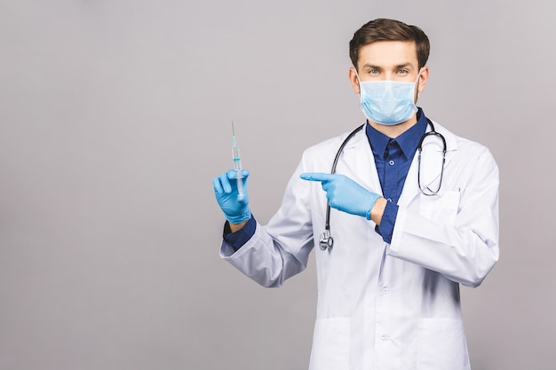 El médico de pie que levanta una inyección pone la máscara en la boca y cuelga el estetoscopio alrededor del cuello con un uniforme blanco, aislado en un fondo gris.