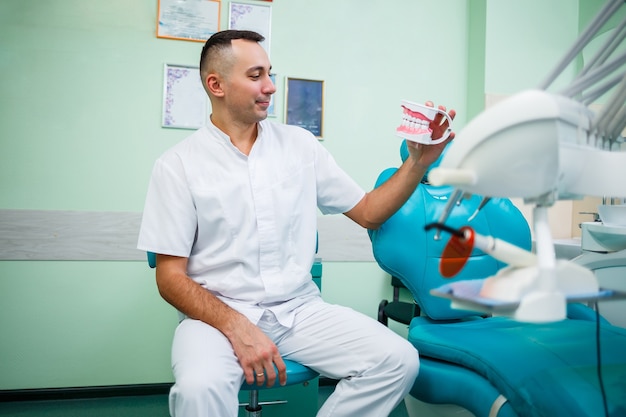 Médico positivo em uniforme branco mostrando mandíbula falsa com dentes enquanto está sentado no consultório odontológico