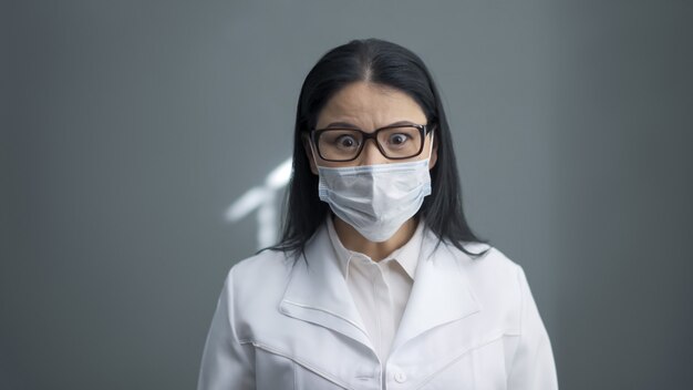 Médico preocupado parece câmera. jovem excitada surpresa em uma máscara protetora e uniforme branco sobre um fundo cinza