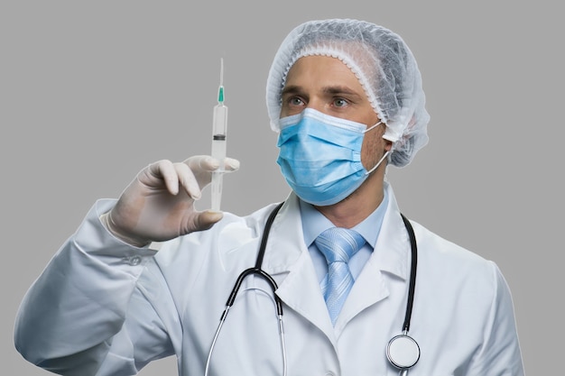 Médico preparando a seringa para injeção. Doutor em máscara facial e chapéu segurando uma seringa em fundo cinza. Prevenção, imunização e tratamento de gripes, vírus e infecções.
