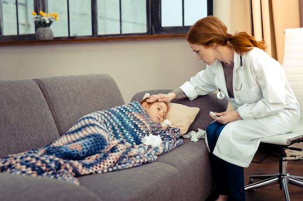 Médico profesional. Agradable joven tocando la frente de la niña mientras controla la temperatura de su cuerpo