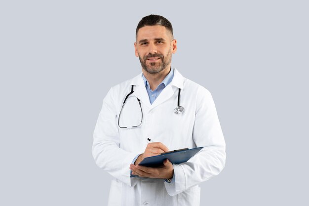 Médico profesional en bata médica con portapapeles en las manos posando sobre una pared gris claro