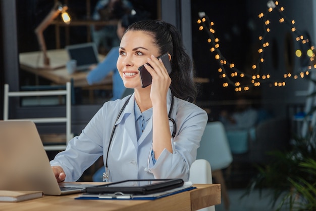 Médico profesional. Hermosa mujer positiva inteligente usando su computadora portátil y hablando por teléfono mientras trabajaba en su oficina