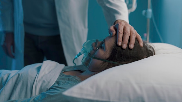 Médico que asiste a una paciente hospitalizada por la noche y le toca la frente. Está acostada en la cama con una máscara de oxígeno.