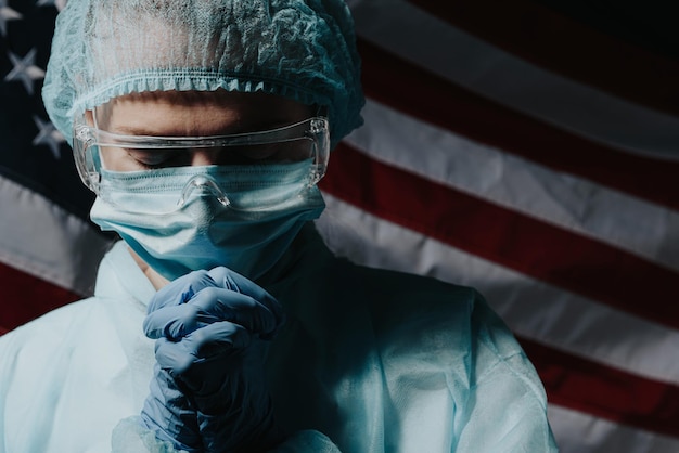Un médico que está cansado después de un duro día, el médico reza con el telón de fondo de la bandera estadounidense y le pide a Dios ayuda y fuerza en la lucha contra la epidemia de coronavirus.
