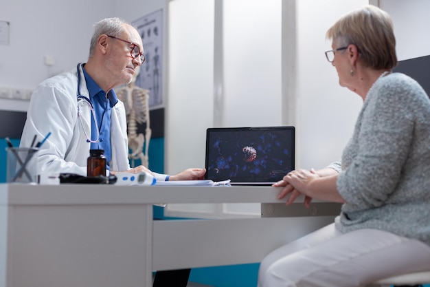 Médico que muestra la ilustración de la bacteria del coronavirus en la computadora portátil al paciente en el gabinete. Médico especialista y paciente mirando la pantalla de la computadora con representación visual de la pandemia de covid 19.