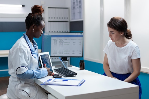 Médico radiólogo afroamericano con tableta que muestra radiografía de huesos a mujer paciente discutiendo síntomas de la enfermedad durante la cita clínica en la oficina del hospital. Radiología corporal en scre