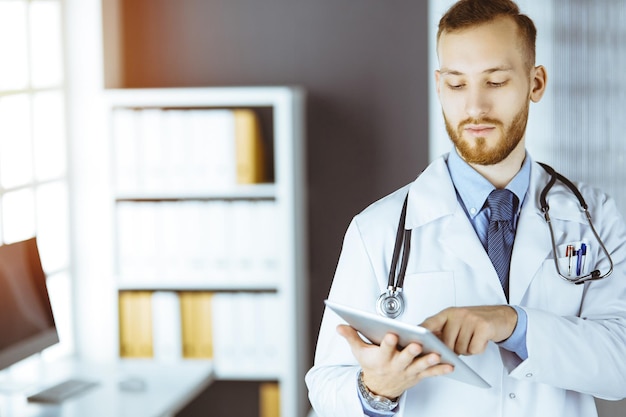 Médico ruivo amigável usando computador tablet em clínica ensolarada em seu local de trabalho Conceito de medicina