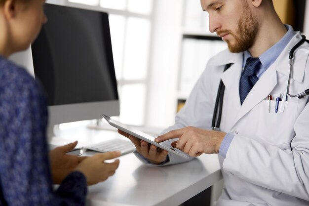 Médico ruivo desconhecido e mulher paciente discutindo o exame de saúde atual enquanto está sentado e usando o computador tablet na clínica, close-up. Conceito de medicina.