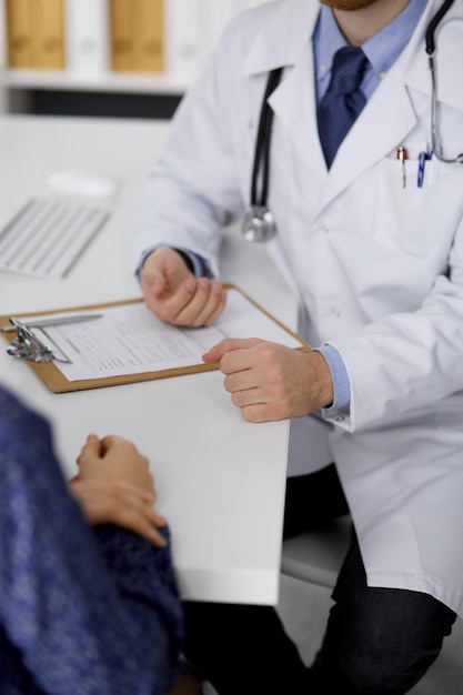 Médico ruivo desconhecido e mulher paciente discutindo o exame de saúde atual enquanto está sentado na clínica ensolarada, close-up. Conceito de medicina.