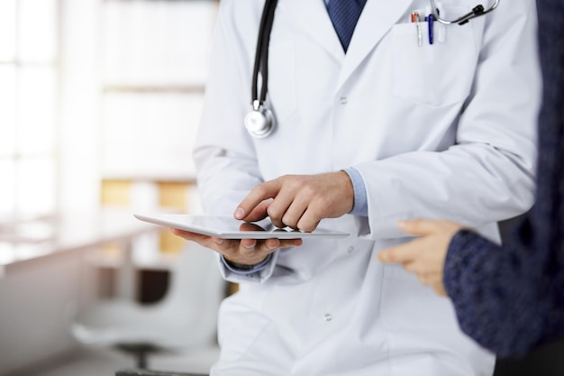 Médico ruivo desconhecido e paciente-mulher discutindo o exame de saúde atual enquanto está sentado e usando o computador tablet na clínica ensolarada, close-up. Conceito de medicina.