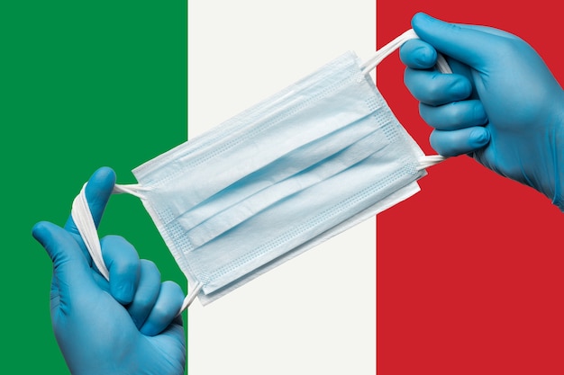 Médico segurando máscara respiratória nas mãos em luvas azuis na bandeira de fundo da itália ou italiano tricolor. quarentena de coronavírus de conceito e surto de pandemia. atadura médica para rosto humano.