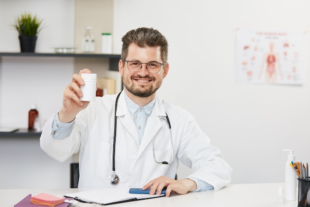Médico segurando o frasco de comprimidos e sorrindo para a câmera, terapeuta barbudo em jaleco branco