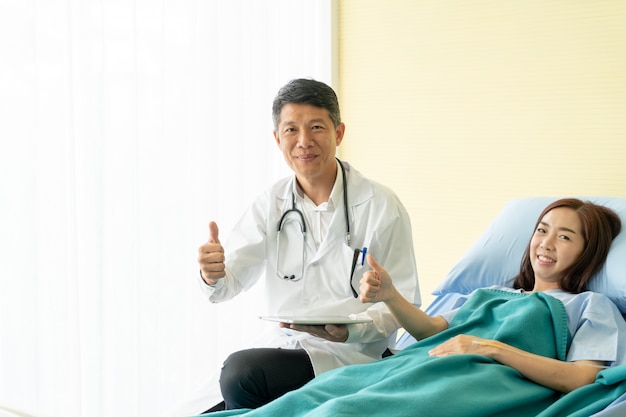 Médico senior asiático sentado en la cama de un hospital y discutiendo con paciente femenino