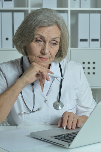 Médico sênior atencioso sentado à mesa com o laptop no escritório