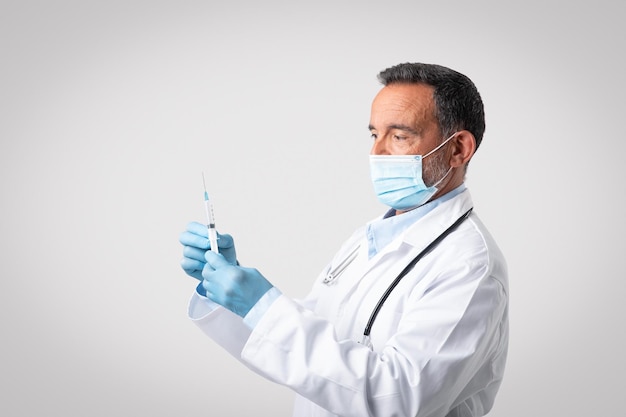 Médico sênior caucasiano sério com máscara protetora de jaleco branco e luvas segura seringa
